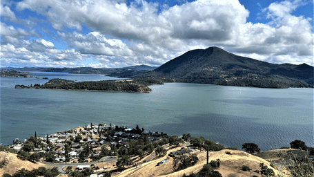 A scenic aerial view of the Clear Lake in Northern California with a small settlement by the lake shores, under a bright blue sky with clouds.