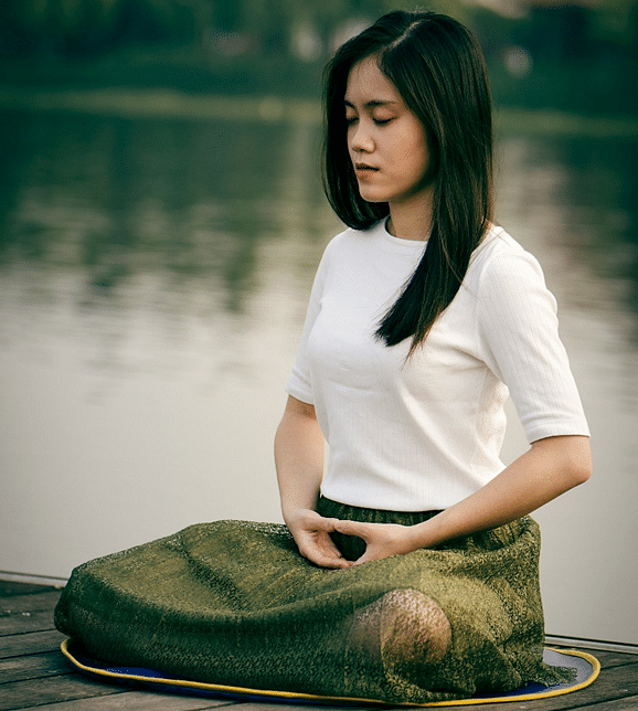 A woman with closed eyes meditates outdoors on a wooden surface, wearing a shirt and skirt