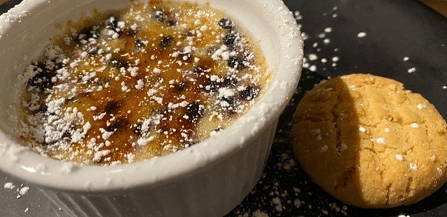 A small casserole dish of crème brûlée with a burnt sugar crust, served with a small cookie.