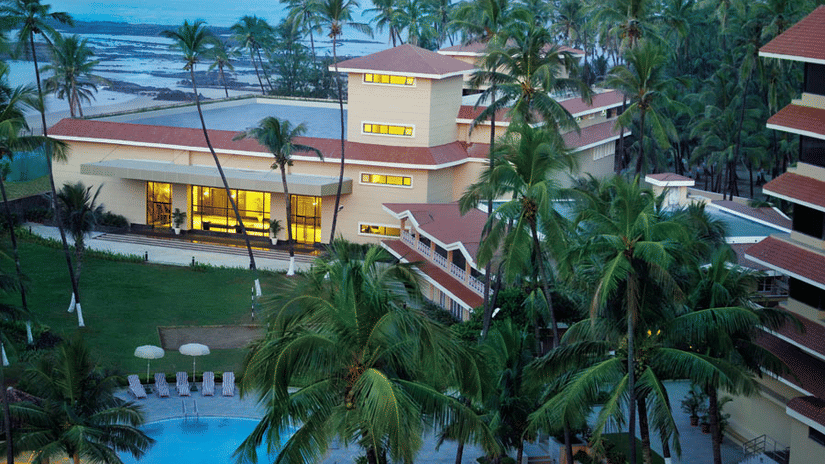 Aerial dusk view of The Retreat Mumbai beachfront resort with warm lit buildings surrounded by tall palm trees and a blue swimming pool in the foreground with the sea visible in the background.