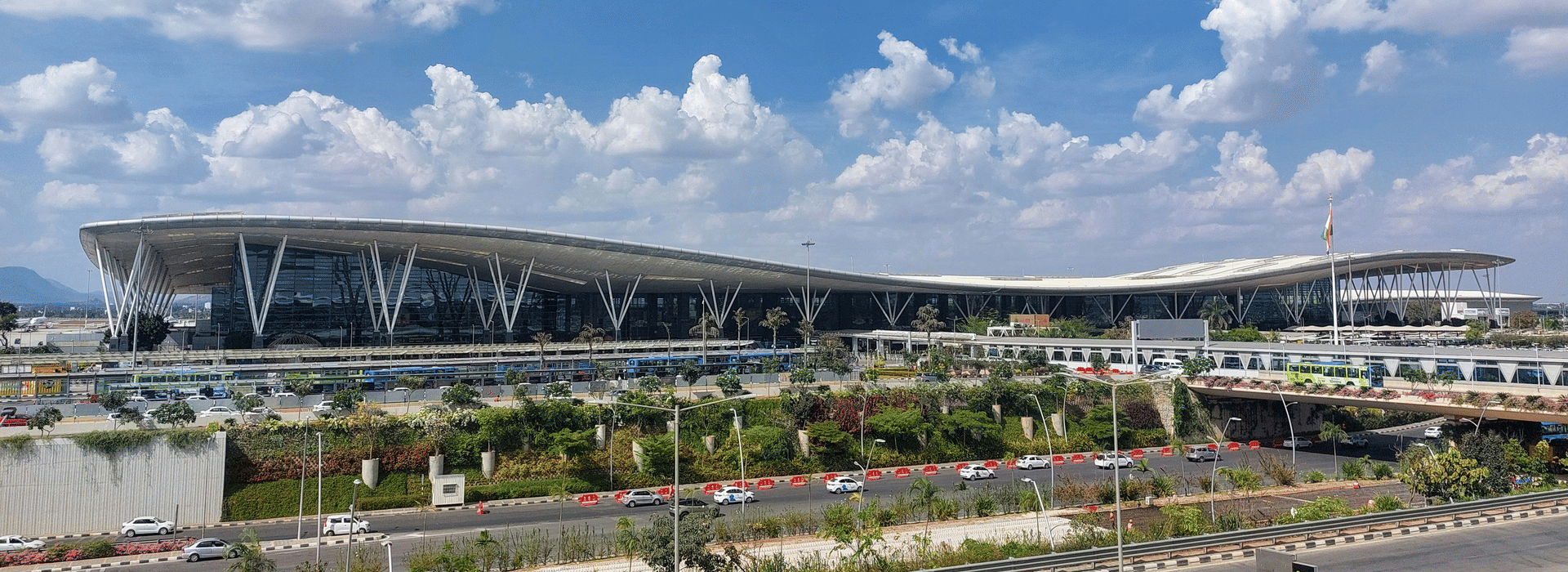 Kempegowda International Airport terminal in Bengaluru with its sweeping curved roof, landscaped approach roads, and a bright blue sky with clouds