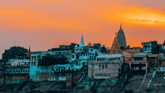 A sunset view of the Varanasi ghats along the Ganges River, featuring buildings and temples with orange hued sky in the background.