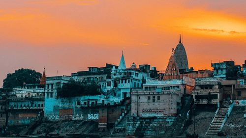 A sunset view of the Varanasi ghats along the Ganges River, featuring buildings and temples with orange hued sky in the background.