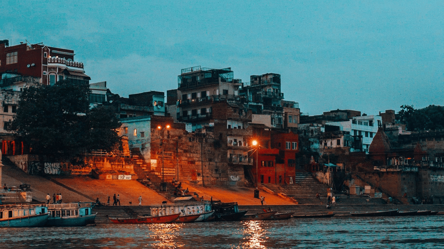 Wide shot of waterfront of Varanasi, featuring illuminated buildings along the Ganges River at twilight.