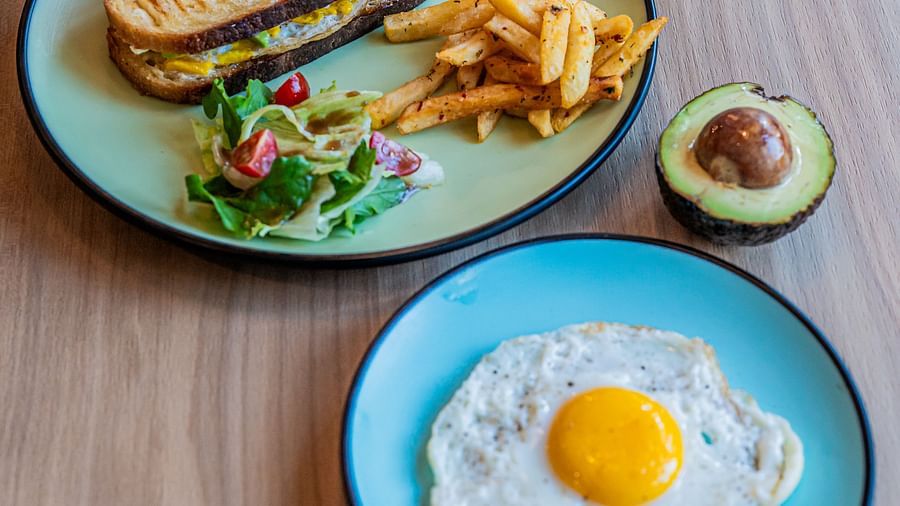 One plate featuring a sandwich with veggies on the side and crispy French fries, while another plate holds an omelette with a half avocado placed beside both plates at Indie Stays Mumbai International Airport.