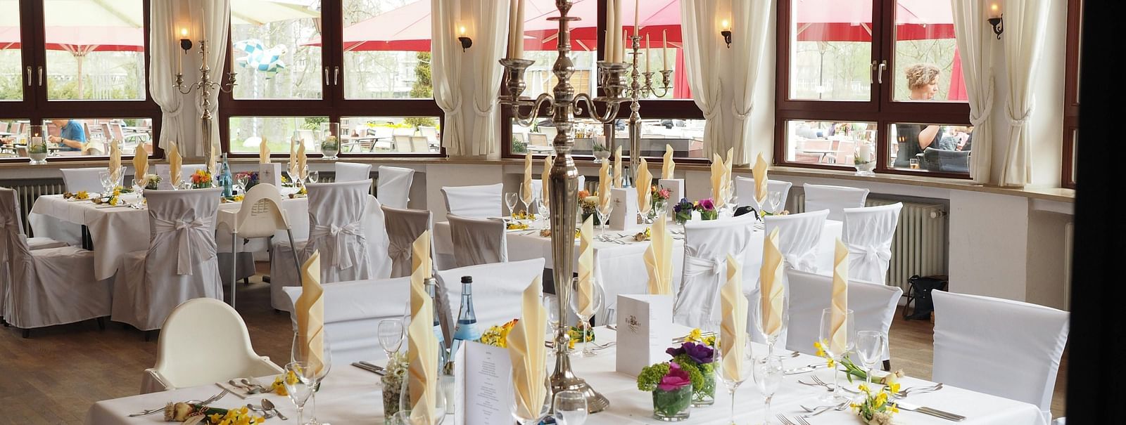 A banquet hall set up with multiple tables draped in white linens and decorated with floral centrepieces, candles, and elegant place settings