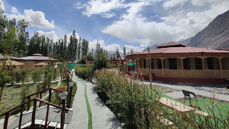 A scenic view of White Sands Resort, Ladakh featuring traditional buildings, lush greenery, and a clear sky with clouds.