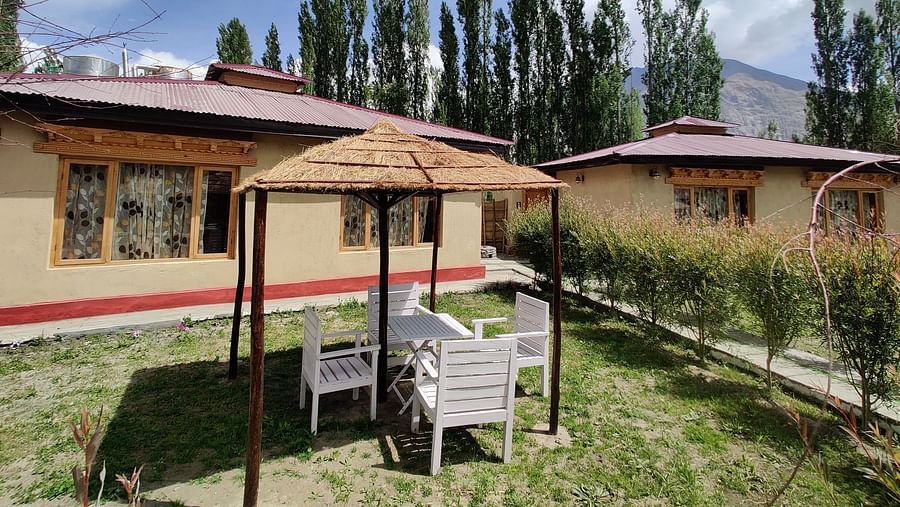 An outdoor seating area of an accommodation building at White Sands Resort, Ladakh, with chairs and a shaded structure, surrounded by trees and greenery, under a partly cloudy sky.