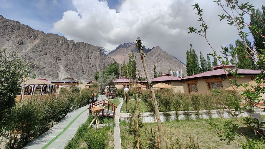 A pathway leading through White Sands Resort, Ladakh, with accommodation buildings and trees on either side, set against a backdrop of mountains and a partly cloudy sky.