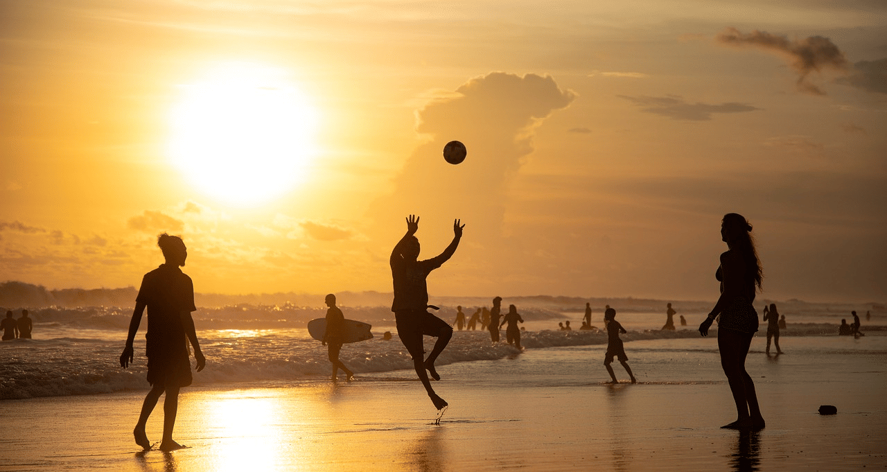 A group of people playing volleyball on a beach at sunset, with the sun reflecting off the wet sand.