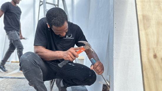 A carpenter in a black shirt uses a hammer and chisel to install a door hinge outdoors.
