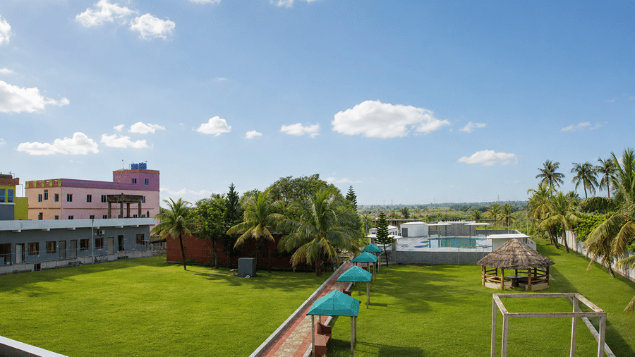 Aerial view of the grounds at Pride Elite Mandarmani showing the lawn, a thatched gazebo, the rooftop pool, and surrounding buildings and palms.