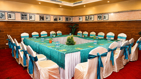 Boardroom set up with a U-shaped table dressed in teal cloth, white chair covers with teal sashes, a red carpet and framed photos on the wall.