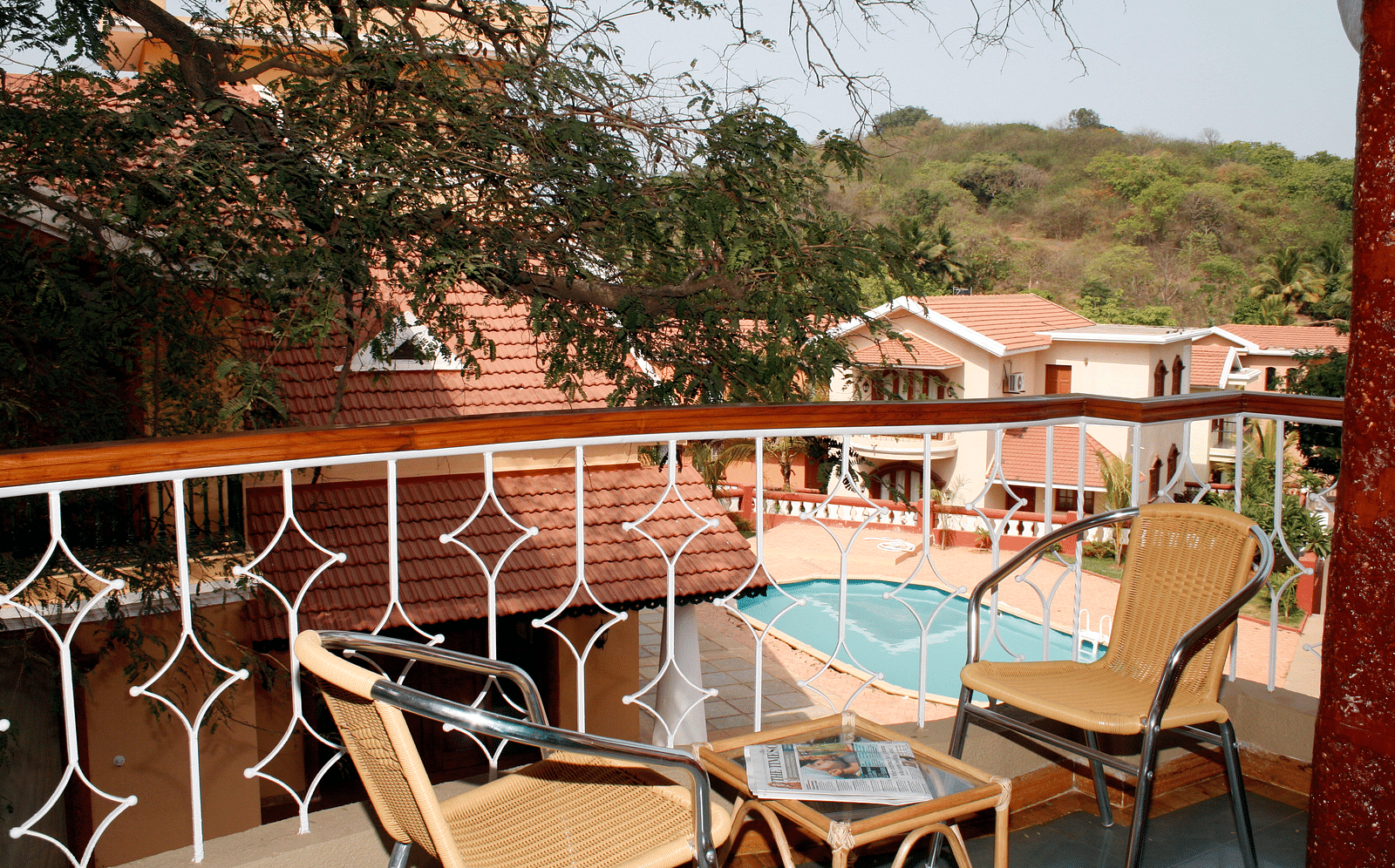 two chairs and a coffee table on the balcony with a view of the greenery in distance and the pool inside the premises