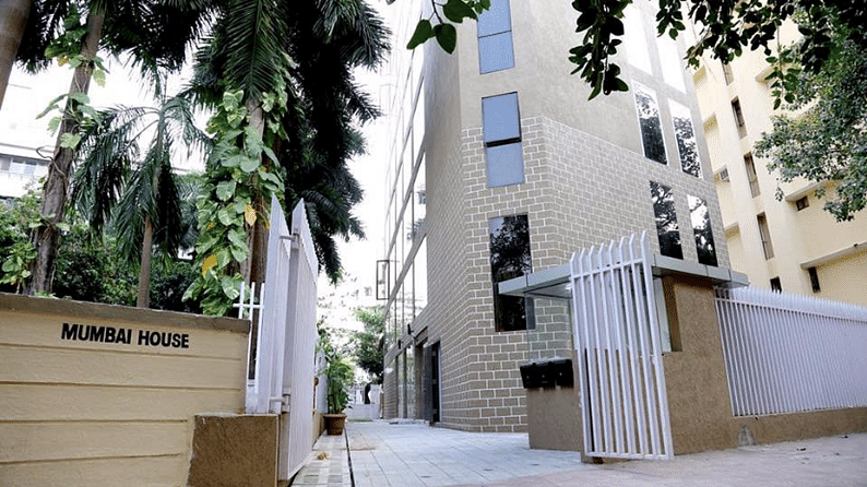 A modern multi-story building labeled MUMBAI HOUSE with a brick facade and reflective windows, flanked by lush green palm trees and a metal gate along a paved pathway.