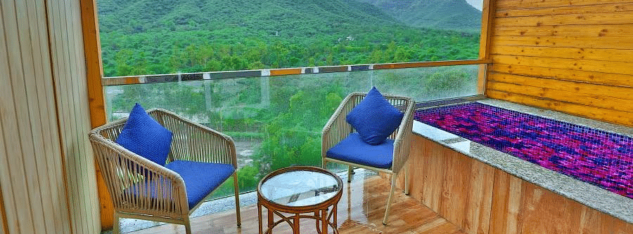 A balcony with two chairs, cushions, and a table overlooks a lush green valley with mountains.- Bamboo Saa Resort & Spa, Udaipur