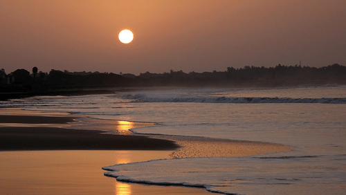 A sunset view over the sea with waves at the shore and mountains in the background.