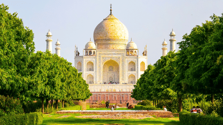 The Taj Mahal viewed from the lush Mughal gardens with tree-lined pathways leading towards the iconic white marble monument.
