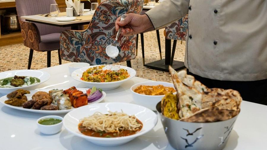 A chef in a beige uniform and black hat plating food at a table filled with Indian dishes in a well-lit restaurant. – Clarks Inn Suites, Near Taj Mahal