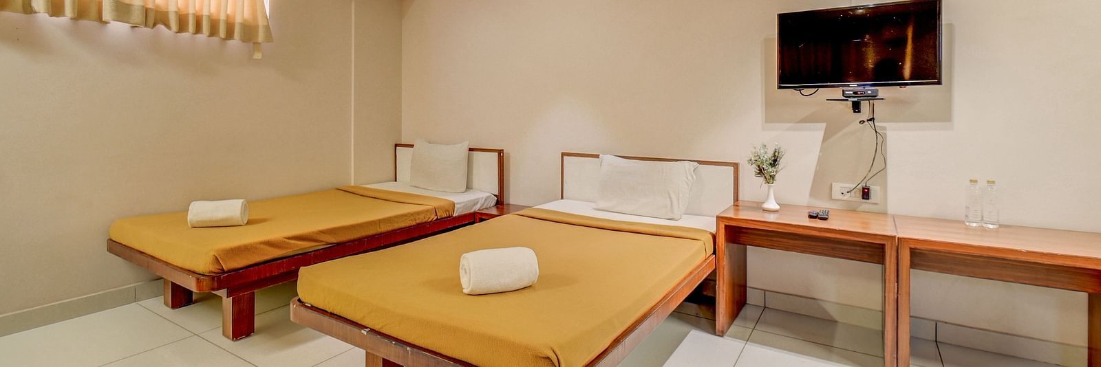 A spacious room at Harigovinda's JNStay, featuring a wall-mounted TV above a wooden table set against the wall, and two beds