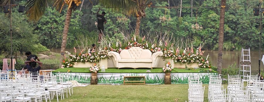 A wedding stage set against a backdrop of palm trees and a river, with rows of white chairs arranged on a lawn.