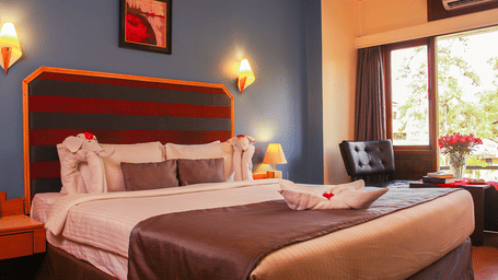 Boutique Room at Polo Towers, Shillong with a king bed, red and grey striped headboard, wall sconces, framed art and a window seat with armchair.