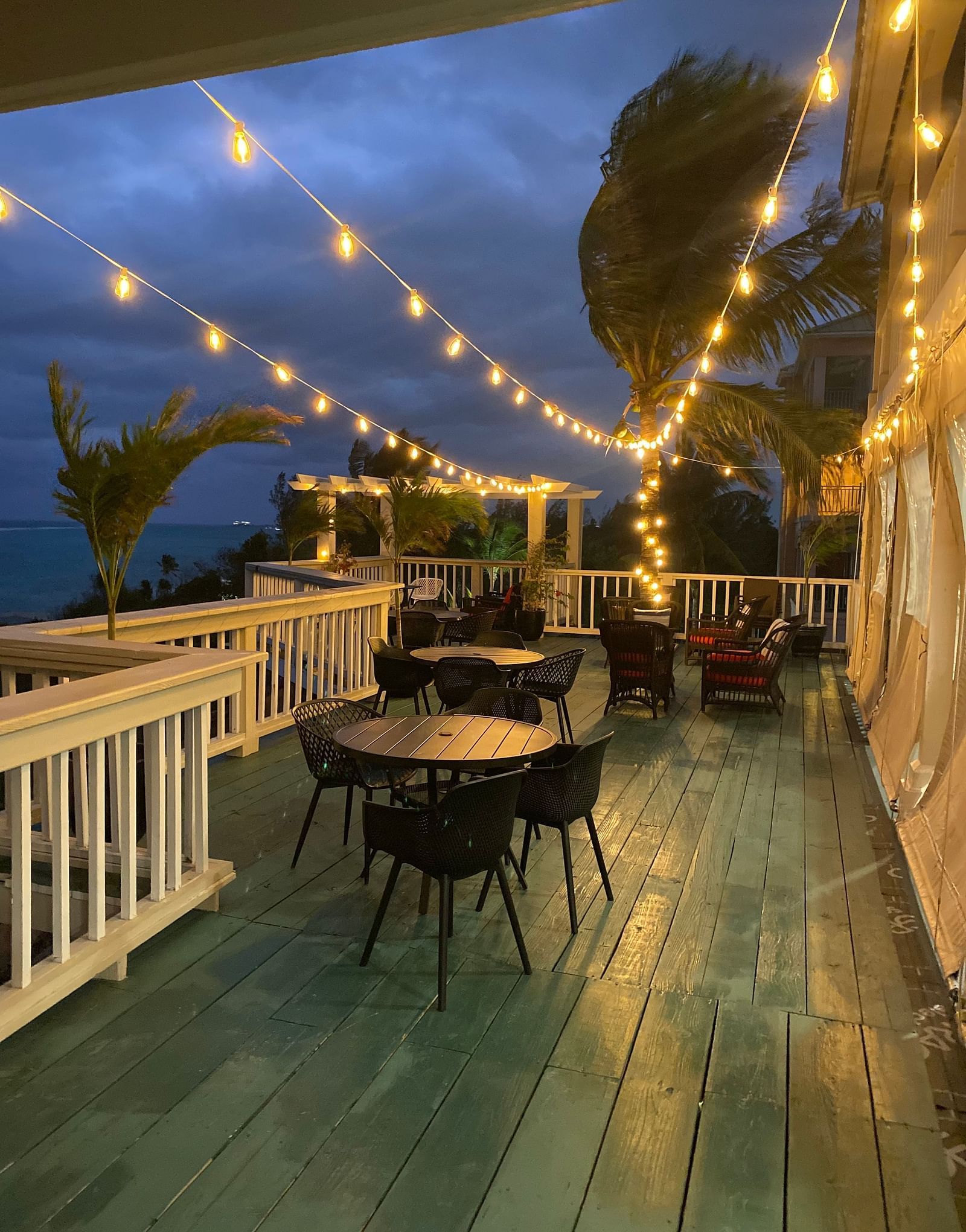 Outdoor dining area with string lights at dusk, white railing and palm trees in view | A Stone's Throw Away
