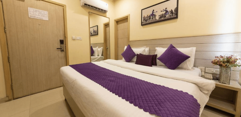 A well-lit hotel room, featuring a bed with white linens and a purple runner and pillows, a light wooden headboard, and a wooden door | Perfectstayz Value Sagar