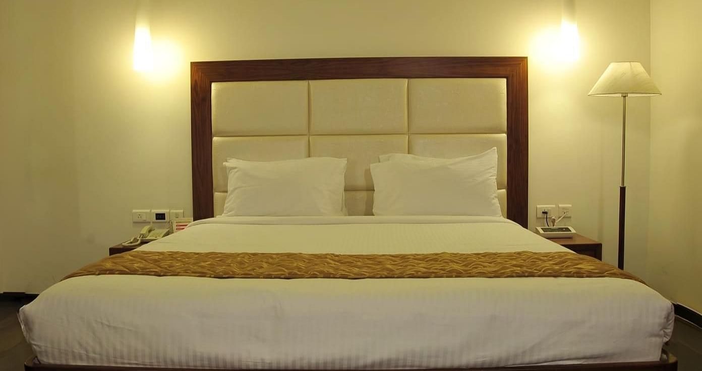 A queen size bed with white linens and a gold runner, two hanging lights, a floor lamp, and a white rug on a wooden floor at Dhanalakshmi Srinivasan Hotels, Perambalur.