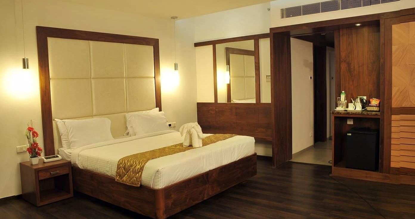 A room with a queen size bed, two side tables, wooden flooring, two mirrors, hanging lights, and a counter with bottles and appliances at Dhanalakshmi Srinivasan Hotels, Perambalur.