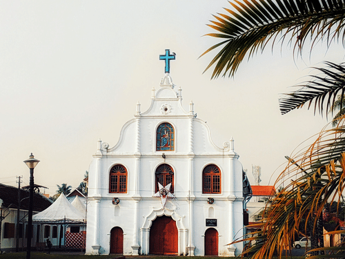 A white church with red doors and a cross stands peacefully beneath a clear sky, framed by tropical palm trees.
