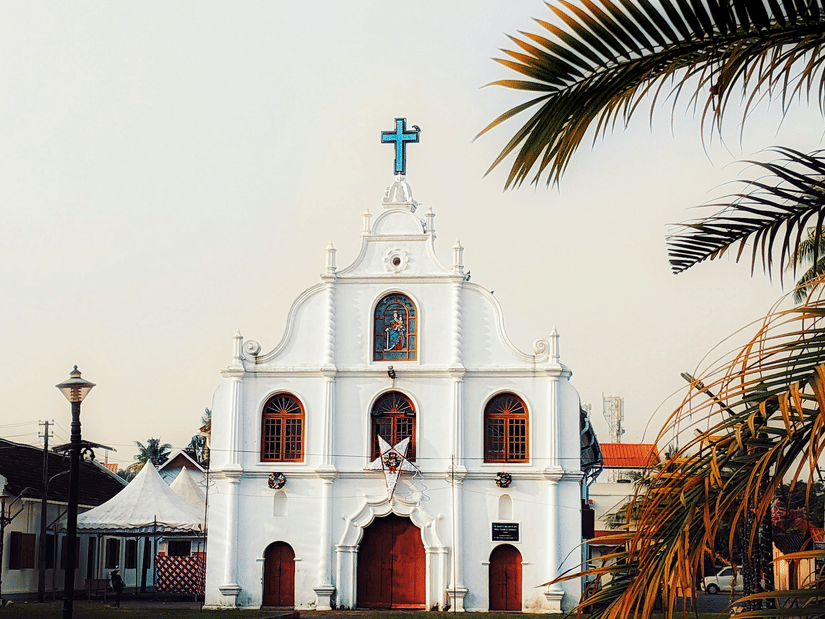 A white church with red doors and a cross stands peacefully beneath a clear sky, framed by tropical palm trees.