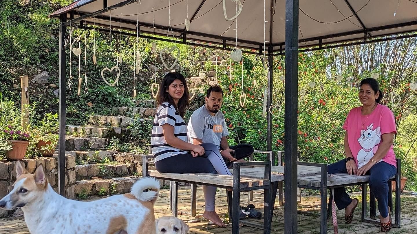 Three people sitting on a shaded bench with a dog watching a view outside in The Sylvan Retreat, Dehradun.