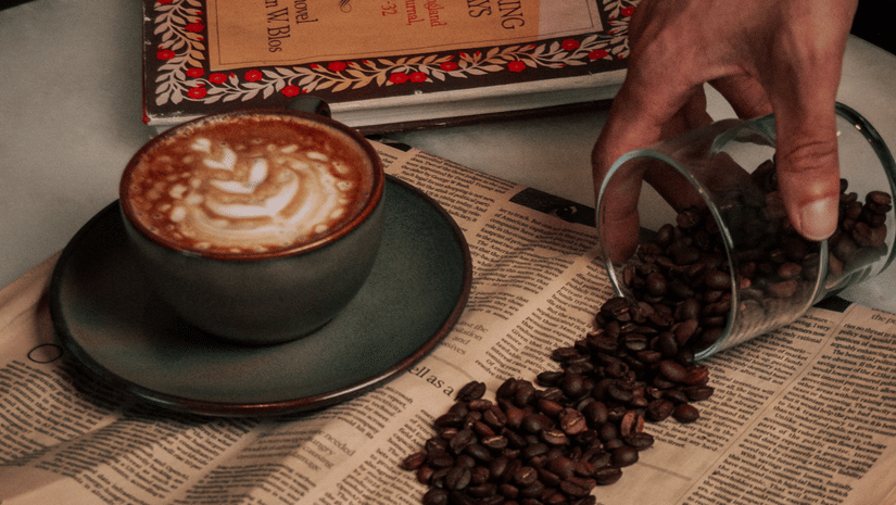 A cup of freshly brewed coffee beside an open book, with roasted coffee beans being poured onto the pages.