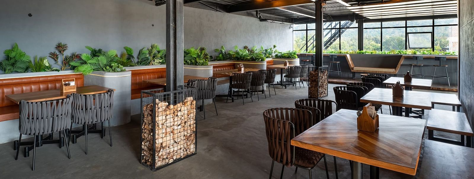 A modern open-air restaurant with wooden tables, cushioned seating, industrial-style ceiling, and lush greenery, offering a stylish dining experience at TGI Livancia Hillside, JC Nagar, Mysore.