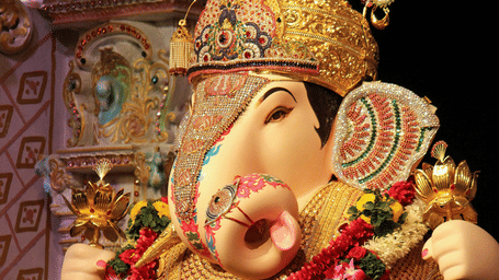 A richly decorated Lord Ganesha idol seated on an ornate silver platform, adorned with an elaborate golden crown, jewellery, floral garlands, and traditional embellishments.