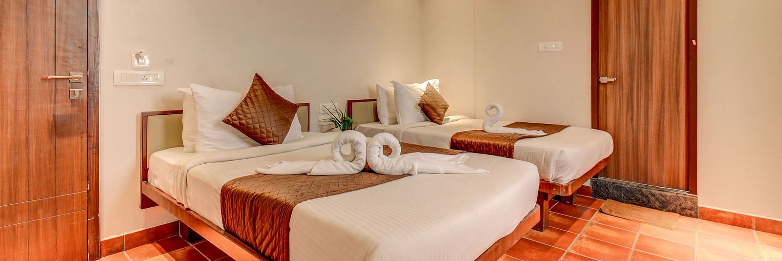 Two neatly made beds placed side by side, with doors on both sides and a wall-mounted AC above one door at Harigovinda's JNStay