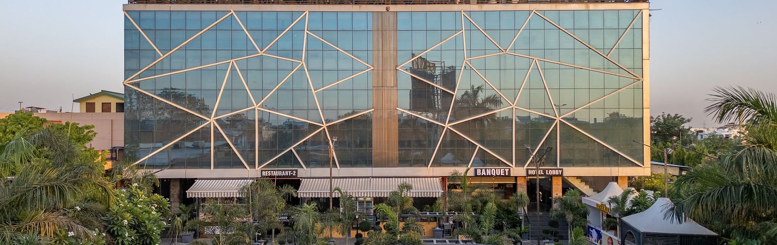 Exterior view of Hotel Highway King, Jaipur, with a large glass facade and rooftop signage, surrounded by greenery.