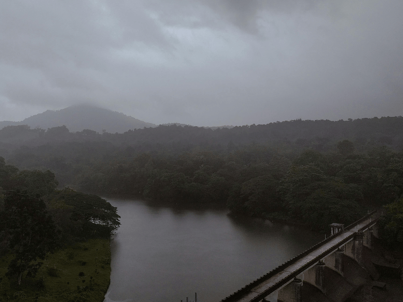 A large concrete dam stretching across a valley between green hills under a cloudy sky.