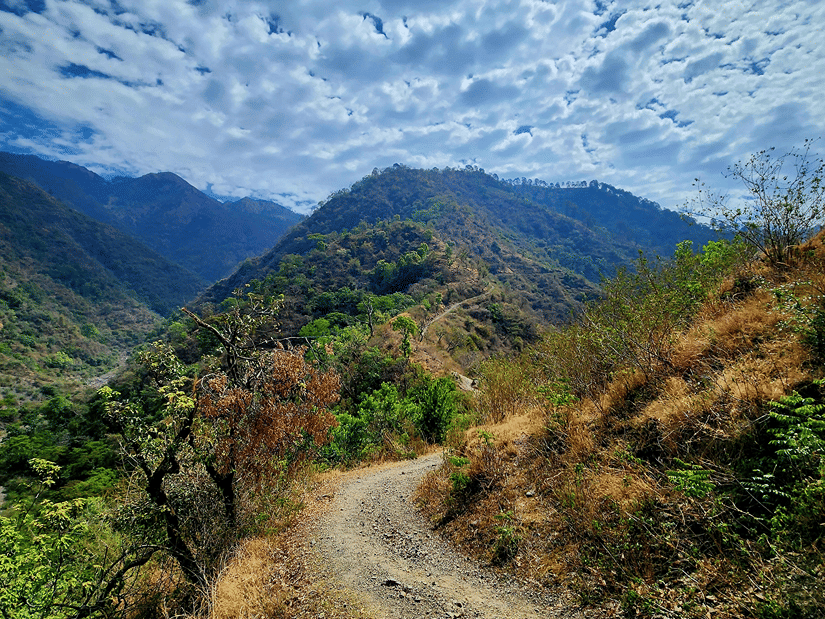A winding mountain road during trekking near Dehradun cuts through rugged Himalayan hills beneath a dramatic sky filled with white clouds and blue patches