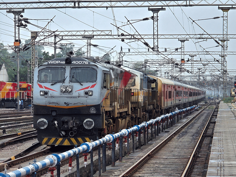 A passenger train travelling along railway tracks beneath overhead electric wires, with railway platforms and infrastructure alongside.