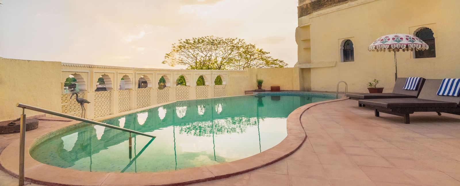 A tranquil pool at Fort Barli, flanked by historic walls and lounge chairs, offers a serene escape with a touch of grandeur.