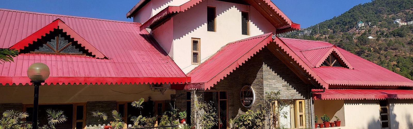 Facade with red rooftops, a landscaped entrance, and a clear blue sky in the background at Dagshai Manor Kasauli - Inde Hotels.