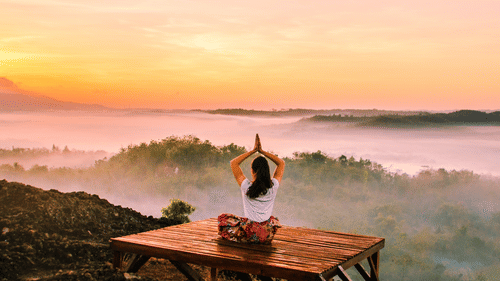A woman doing meditation sitting in open facing green valley and orange hued sky.