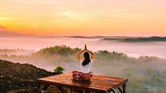 A woman doing meditation sitting in open facing green valley and orange hued sky.