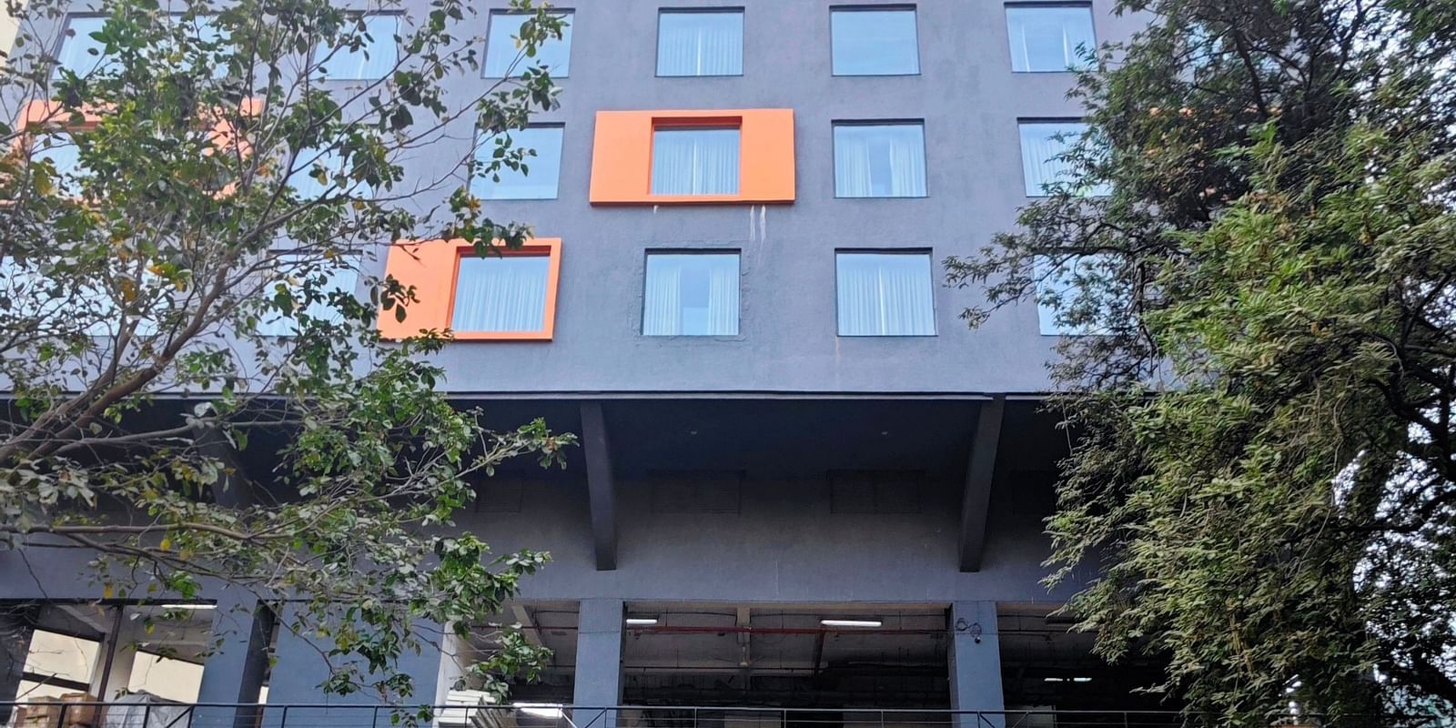 A modern, multi-story hotel building with a dark grey facade and bright orange window accents. The hotel has a covered parking area in front, and there are trees and shrubs surrounding the building.