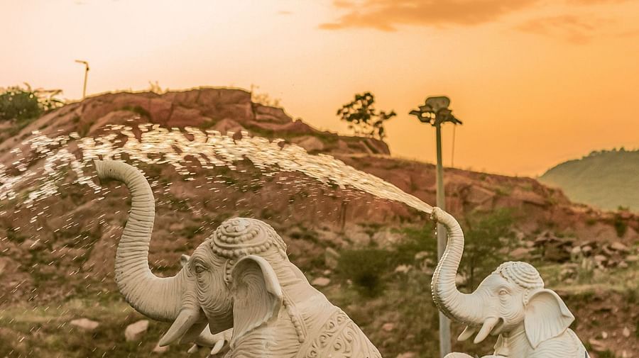 An image of two elephants on a metal structure with water coming from their trunks like a fountain and a small hill in the background
