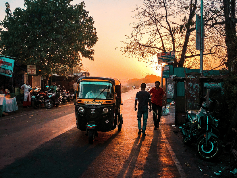 A view of the sun setting in the background with two people standing next to an auto rickshaw and numerous trees on either side of the road