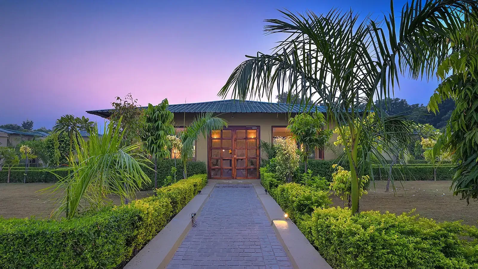 A paved walkway leads to a wooden double-door entrance, surrounded by lush greenery and warm lighting. The villa has a green sloped roof and large windows, set against a twilight sky - Singhvilas, Ranthambore