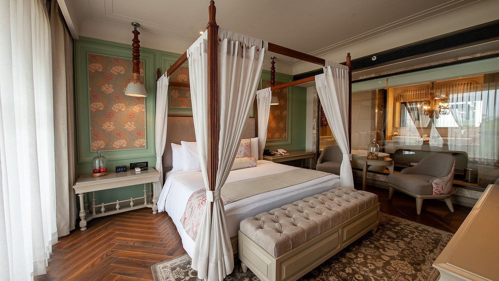 A wide view of the elegant suite at The Evren, Vagator, showcasing a four-poster bed, a stylish rug on a herringbone floor, and a seating area with two armchairs near a window. The room has a view into the connected bathroom.